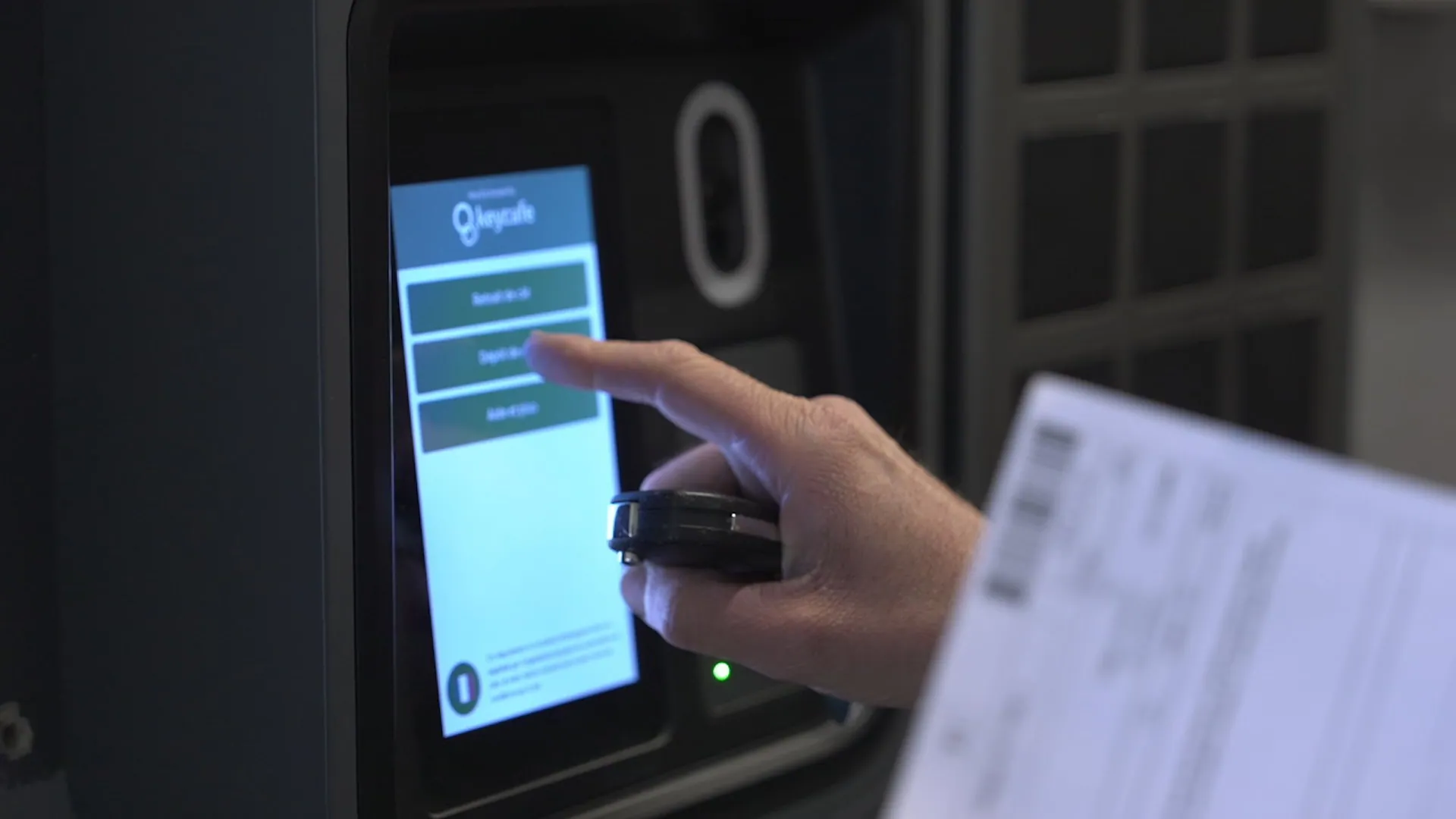 Close-up of a hand touching the Keycafe smart kiosk screen, displaying key management options, while holding a car key fob.