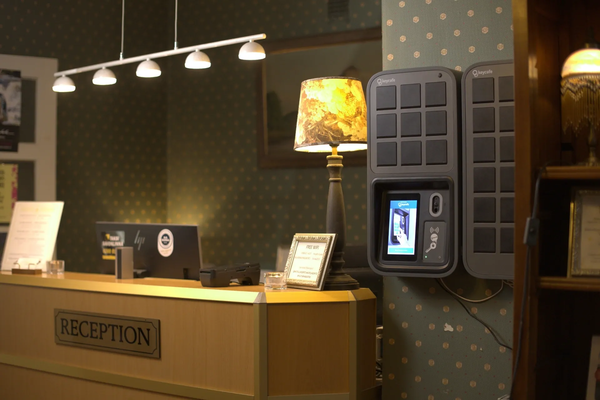 Two Keycafe smart key lockers mounted on a patterned wall next to a wooden hotel reception desk with a lamp and computer.