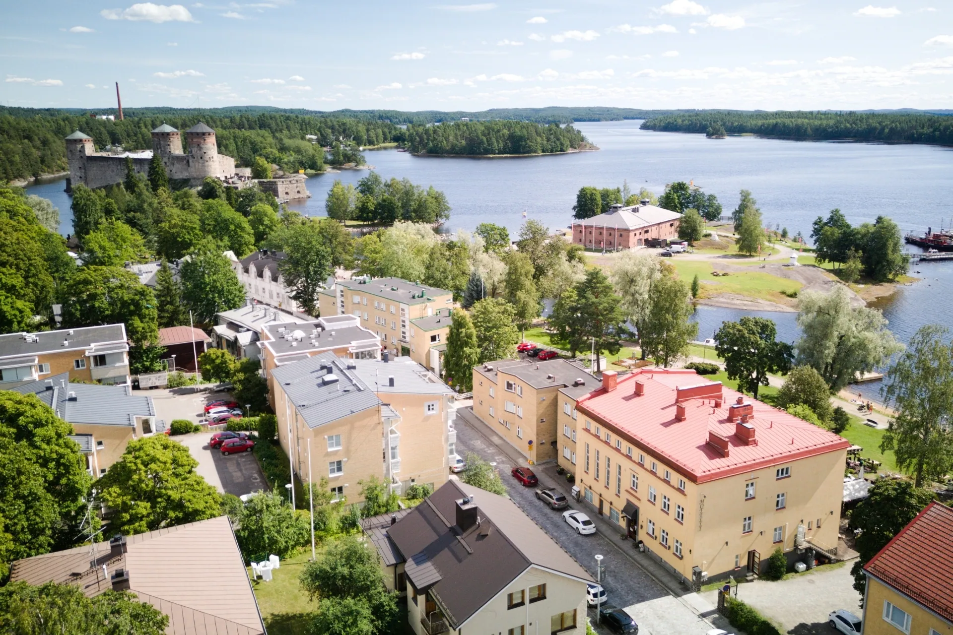 Aerial view of a picturesque lakeside town featuring a medieval castle, residential buildings, and forested islands under a blue sky.