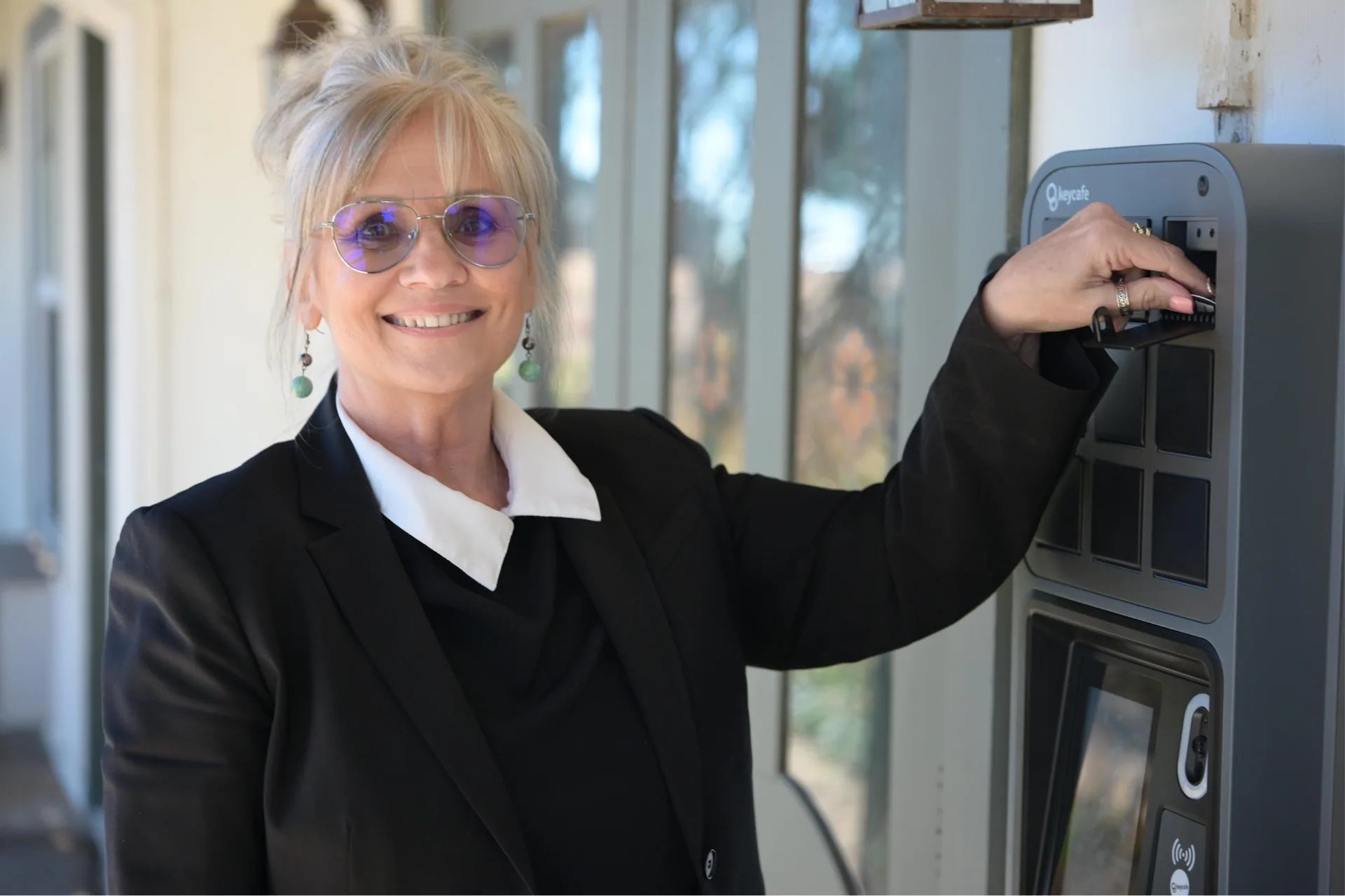 A smiling blonde woman in a black blazer retrieving a key from a Keycafe terminal.