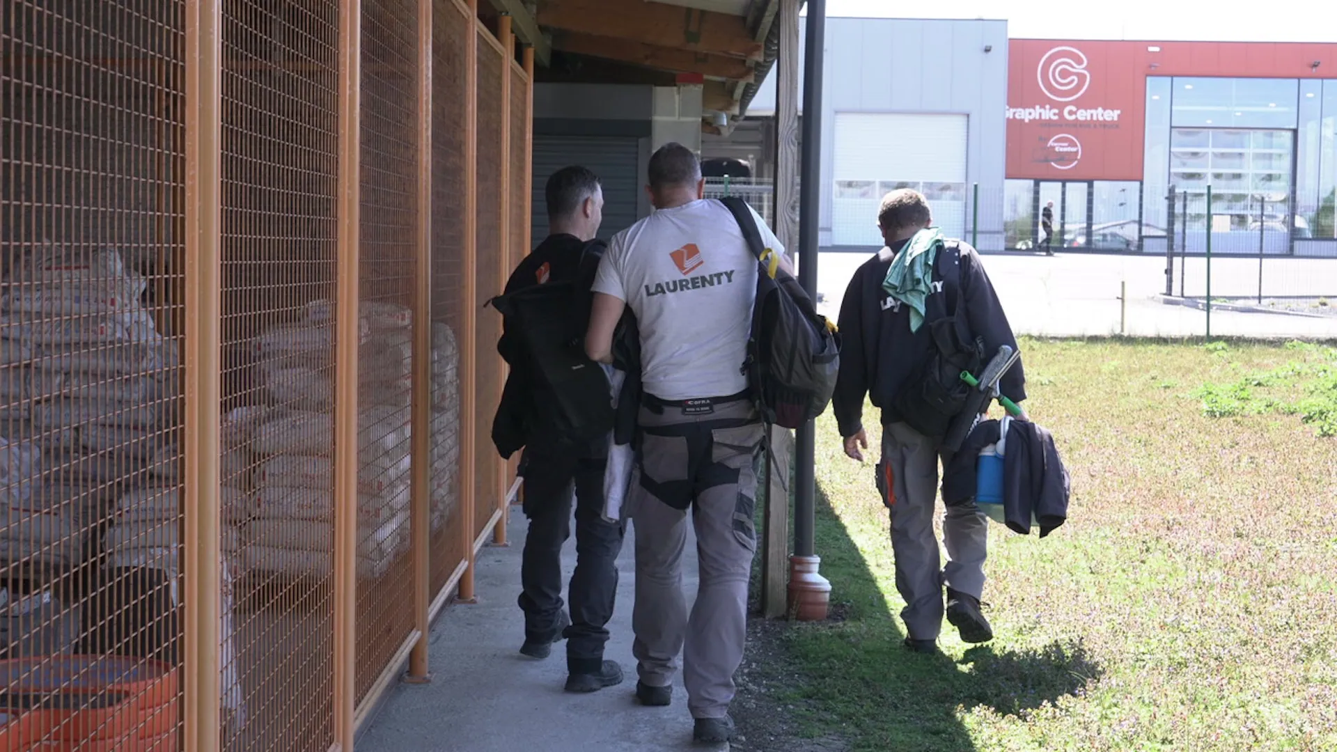 Three workers carrying bags and tools walk past storage cages towards a commercial building labeled 'Graphic Center' on a sunny day.