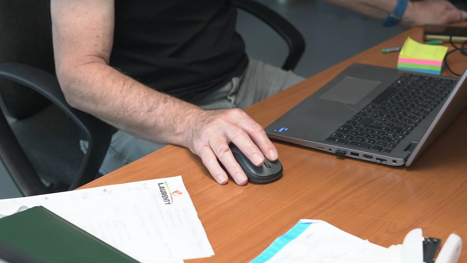 A person's hand uses a computer mouse next to a laptop on a wooden desk with documents and colorful sticky notes.