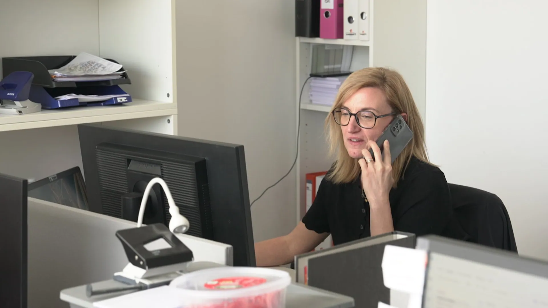 A blonde woman wearing glasses speaks on a smartphone while looking at a computer monitor in a bright office environment.