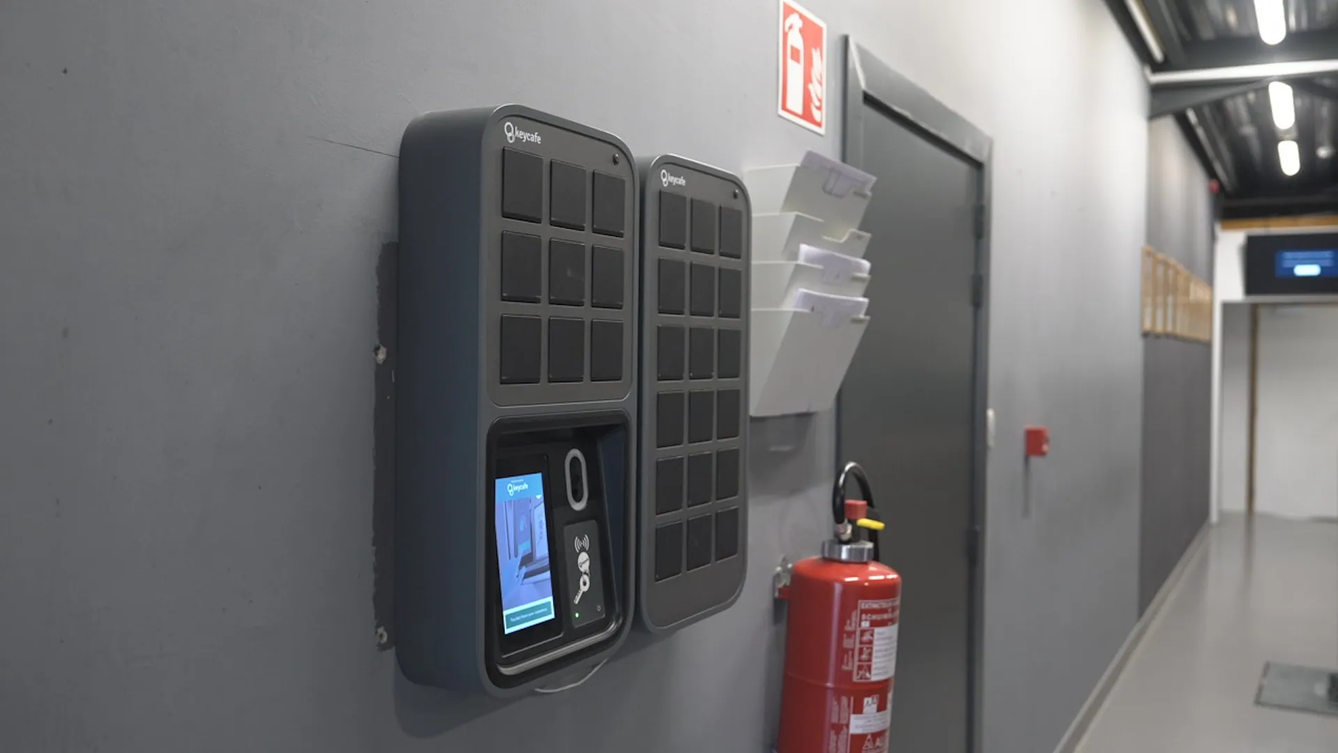 Two Keycafe smart key locker devices are mounted on a gray wall in a hallway, with one displaying the interface. A fire extinguisher and a wall-mounted organizer are nearby.