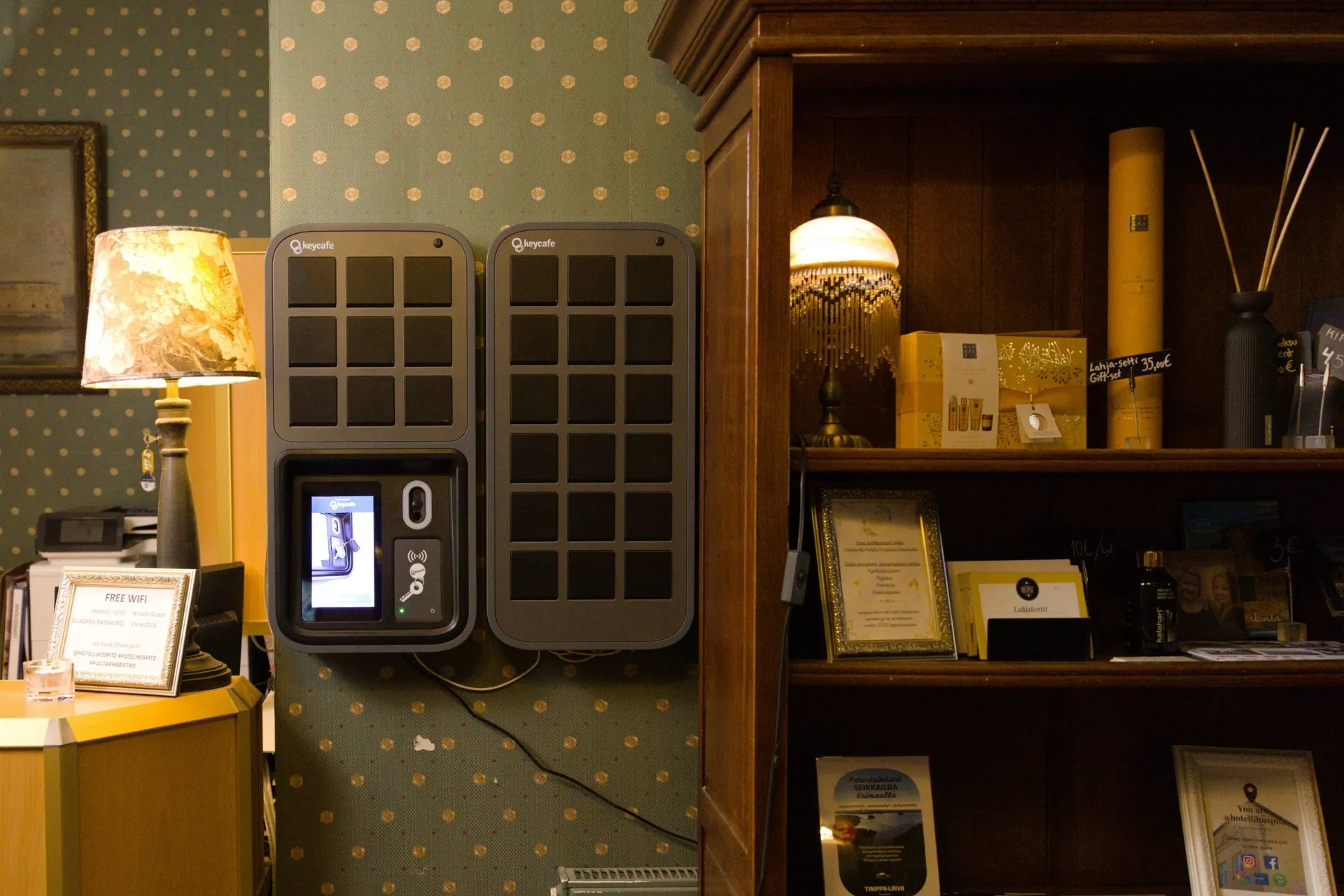 Two Keycafe smart lockers are mounted on a green patterned wall next to a dark wooden bookshelf filled with decor and a table lamp.